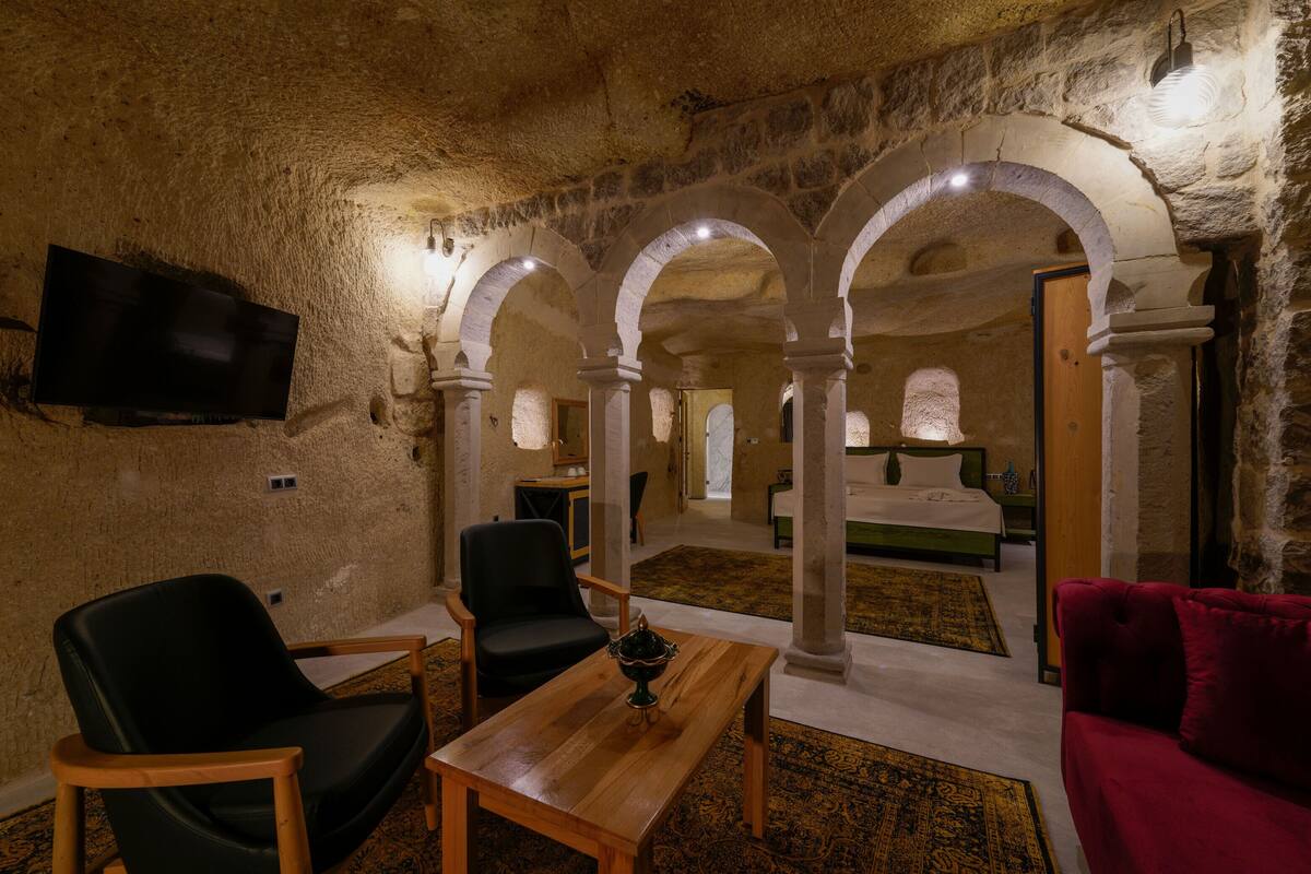 A room with stone walls, a wooden table, two chairs, a red sofa, and a bed with a green headboard.