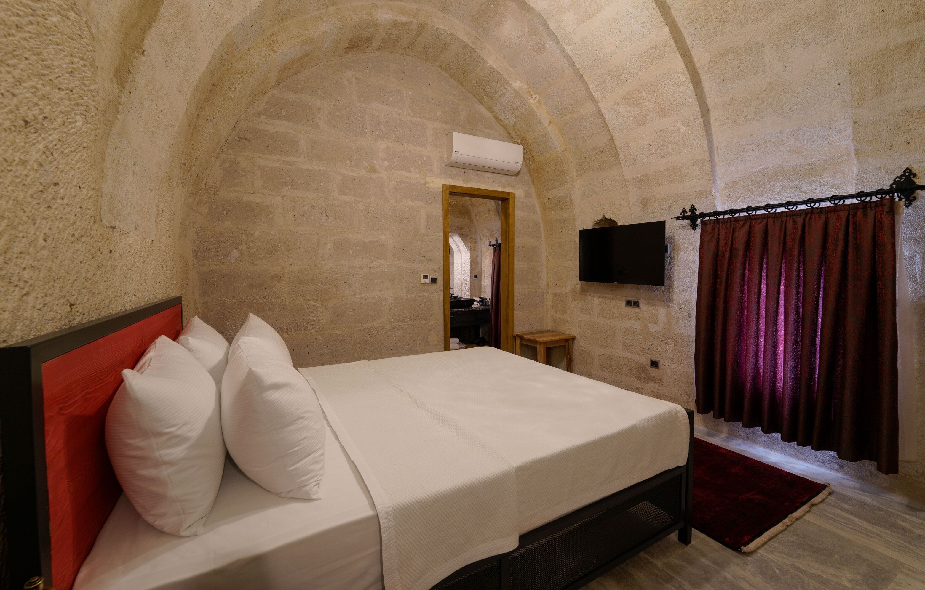 A bedroom with a stone arched ceiling, a bed with white bedding, a small wooden stool, and a TV mounted on the wall.
