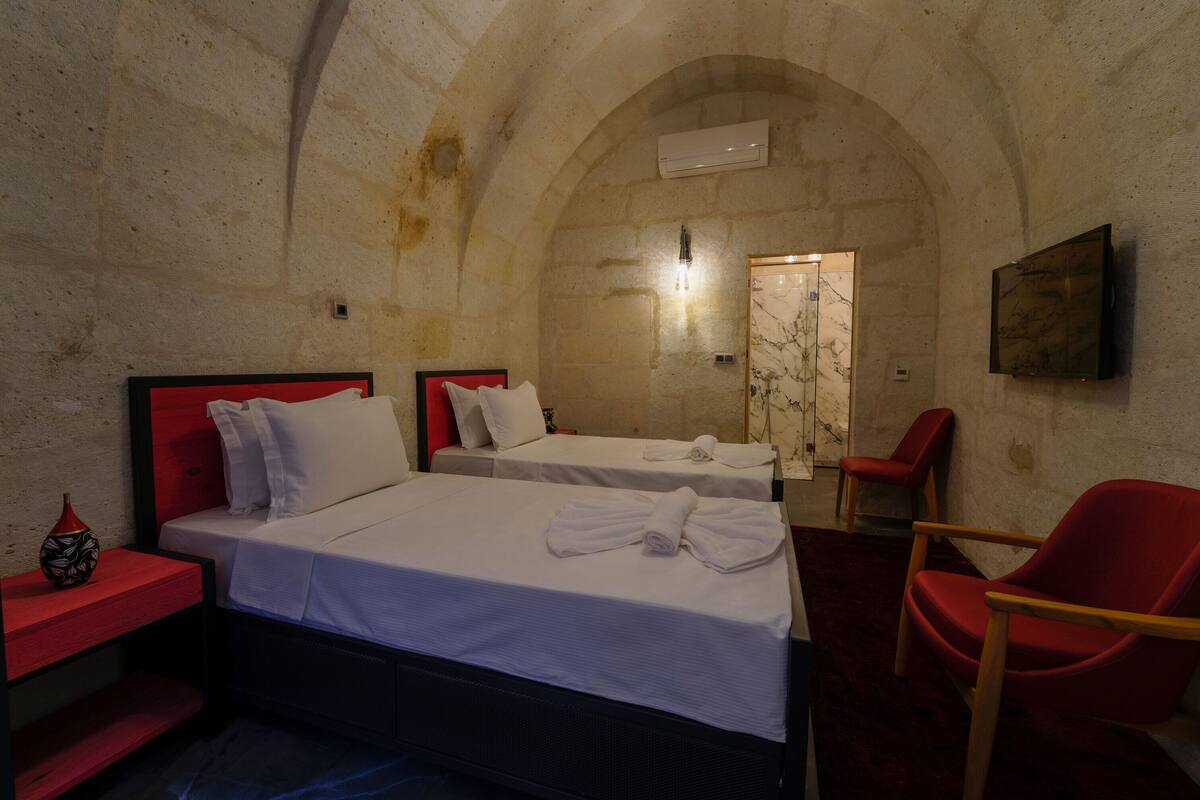 A room with two beds, a red chair, a small table with a vase, and an arched stone wall.
