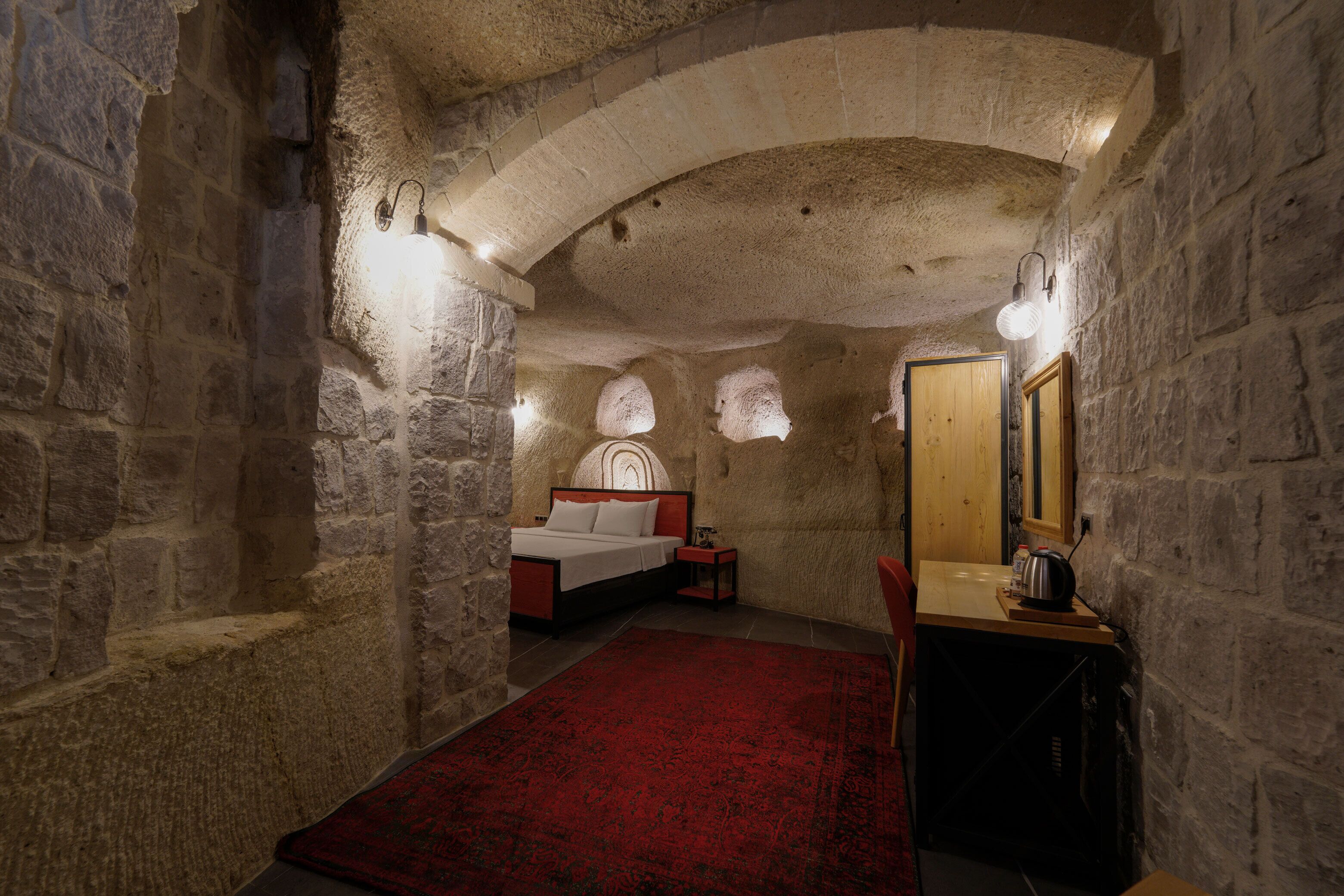 A bedroom with stone walls, a bed, a desk, and a red carpet.