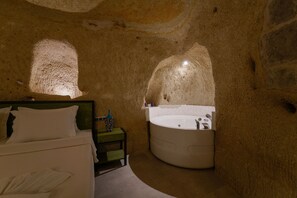 A bedroom with a bed, a nightstand, and a large circular bathtub in a cave-like room with natural rock walls. - The Niche Cave Hotel (Nevsehir)