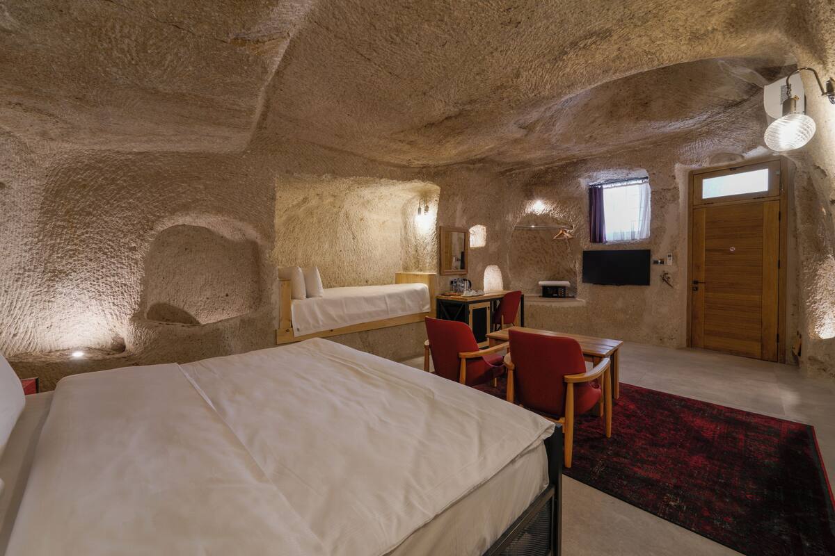 A cozy room with a large bed, a desk, and a chair, featuring a rustic stone ceiling and a wooden door.