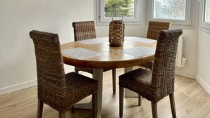 A dining area with a round wooden table, four wicker chairs, a woven pendant light, and a view through white blinds.