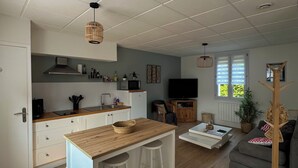 A modern kitchen with a central island, a dining area with stools, a grey sofa, and a TV in a living room.
