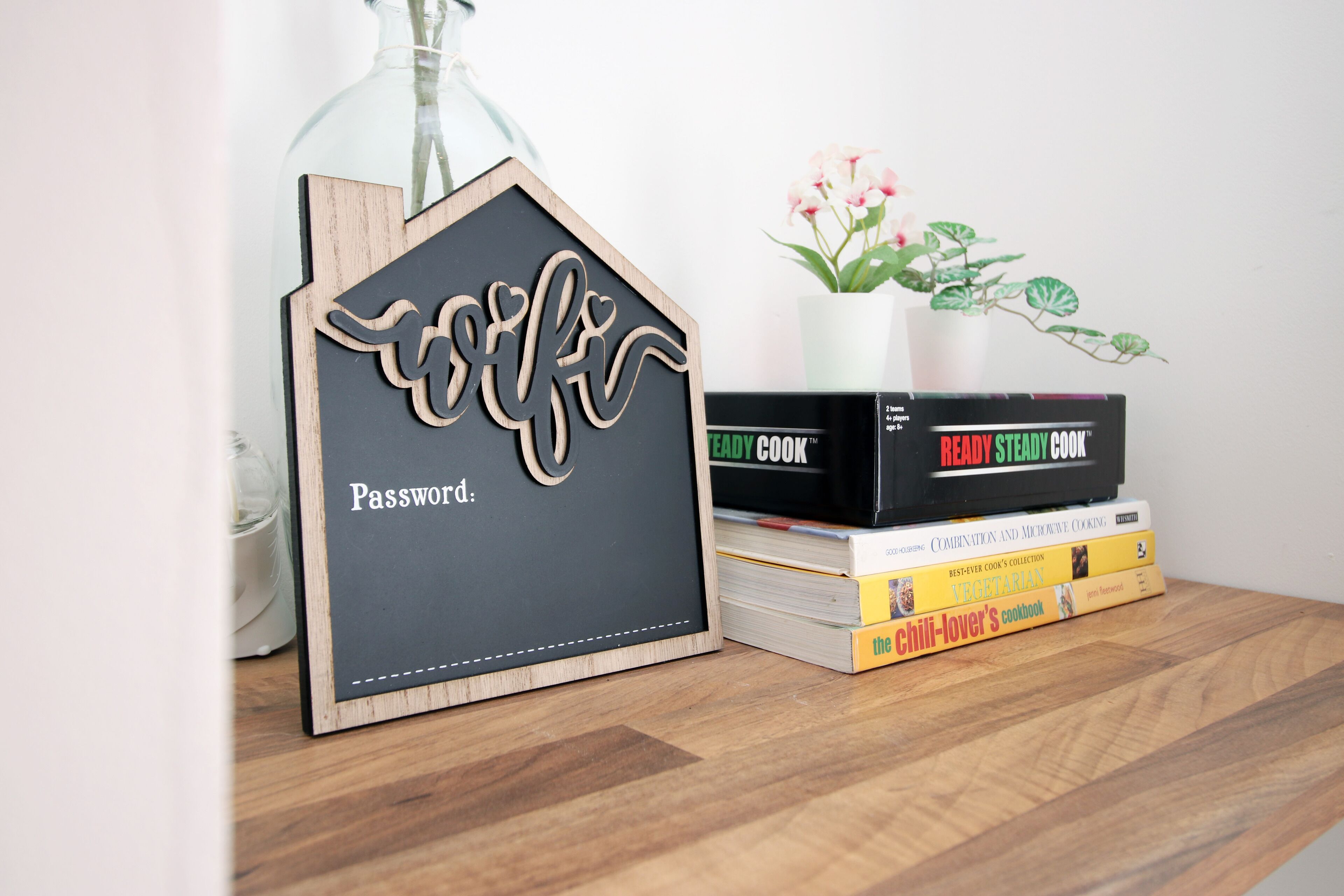 A wooden house-shaped password holder with a plant and cookbooks on a wooden surface.
