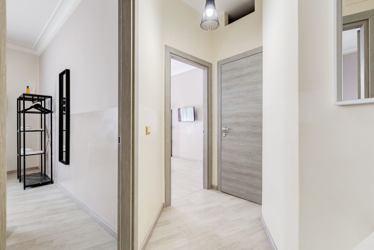 Un pasillo con piso de madera, un estante metálico negro, una puerta con interruptor de luz y una lámpara en el techo.