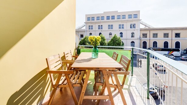 Un balcon avec une table et des chaises en bois, un vase rempli de fleurs et une vue sur un immeuble doté de nombreuses fenêtres.