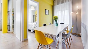Une chambre moderne avec une table à manger blanche, des chaises jaunes, un lit et une grande fenêtre avec des rideaux.