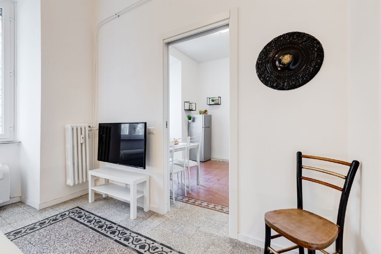 A living room with a TV, a white bench, a patterned rug, and a dining area in the background.