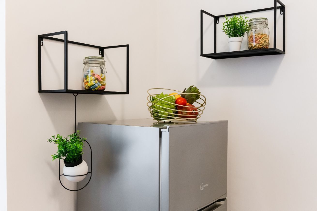 A modern kitchen with a stainless steel refrigerator, two black metal shelves with jars and plants, and a hanging planter with green foliage.