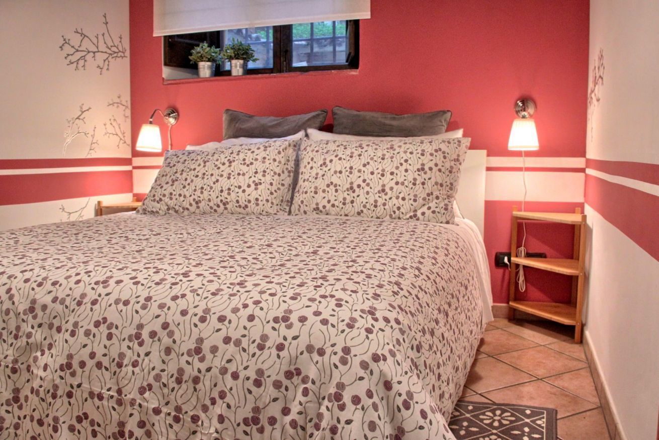 A bedroom with a bed, two bedside lamps, a window with plants, and a red wall.
