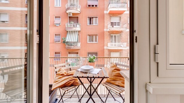 A balcony with a table and chairs, a potted plant, and a view of a multi-story building with balconies.