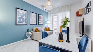 A modern living room with a blue accent wall, a sofa with cushions, a dining table with chairs, a vase with flowers, and framed artwork.