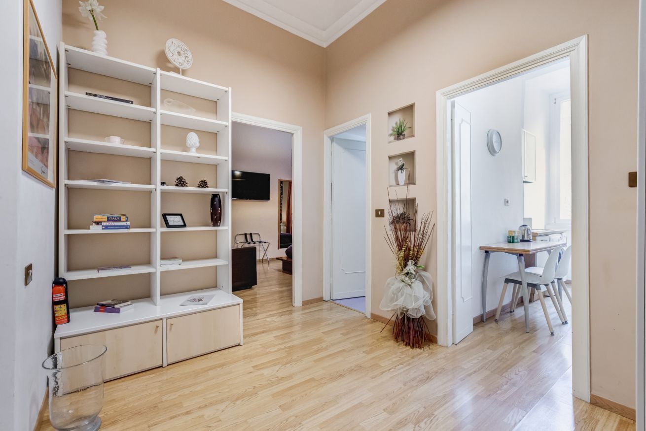Una habitación con piso de madera, una estantería blanca, un comedor con mesa y sillas, y una puerta que da a otra habitación.