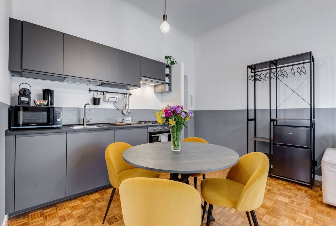 Eine moderne Küche mit grauen Schränken, einem runden Tisch mit gelben Stühlen, einem schwarzen Kühlschrank und einem Kleiderständer.