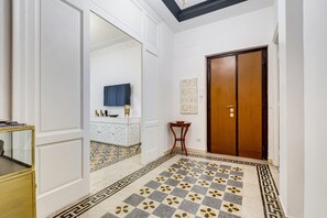 Un corridoio con pavimento a motivi, una porta in legno, una parete bianca con un quadro incorniciato e un tavolino con un vaso.