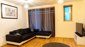 A living room with a black sofa, a small white ottoman, a grey rug, a framed picture on the wall, and a television on a stand. - East London 1-bed Apt, Sleeps 2, With Parking (London)