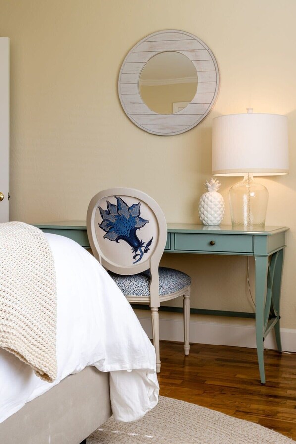 Un dormitorio con cama, escritorio, silla con reposeras decorativas, espejo redondo y lámpara.