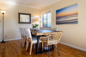 A dining area with a wooden table, wicker chairs, a floor lamp, a mirror, and a painting of a sunset. - Pleasure Point Getaway with hot tub, dog friendly! (Santa Cruz)