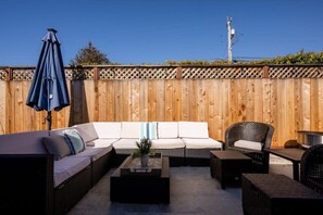 Un patio con cerco de madera, un sofá blanco, muebles de mimbre y una sombrilla azul.