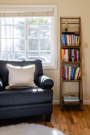 Un salón con un sofá azul oscuro, un cojín blanco, una estantería de madera llena de libros y una ventana con persianas.