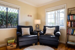 Une pièce comprenant deux fauteuils sombres, une petite table avec une lampe, une bibliothèque et une fenêtre donnant sur un paysage verdoyant.