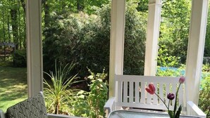 A covered patio with a table and a chair, surrounded by lush greenery. - Kilburn Manor 1820 Historical house (Malone)