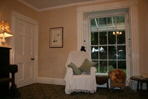 Relax in a cozy room with a large window offering a view of the outdoors, featuring a comfortable seating area with a plush armchair and a charming wicker chair.