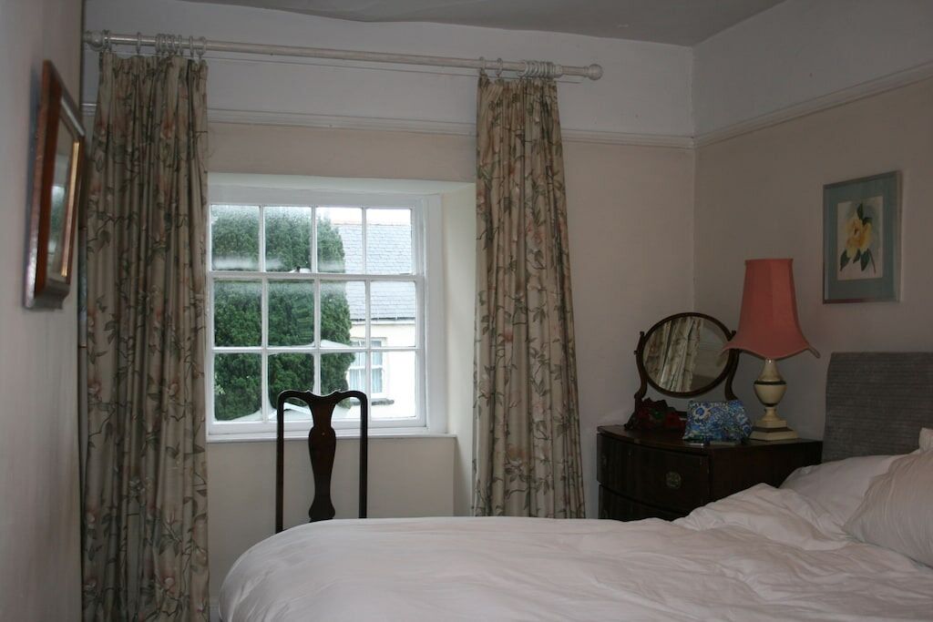 Une chambre à coucher avec un lit, une fenêtre avec des rideaux, une table de chevet avec une lampe et un miroir.