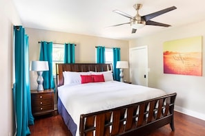Quarto com cama de madeira, cortinas verde-água, ventilador de teto e parede com pintura de pôr do sol.