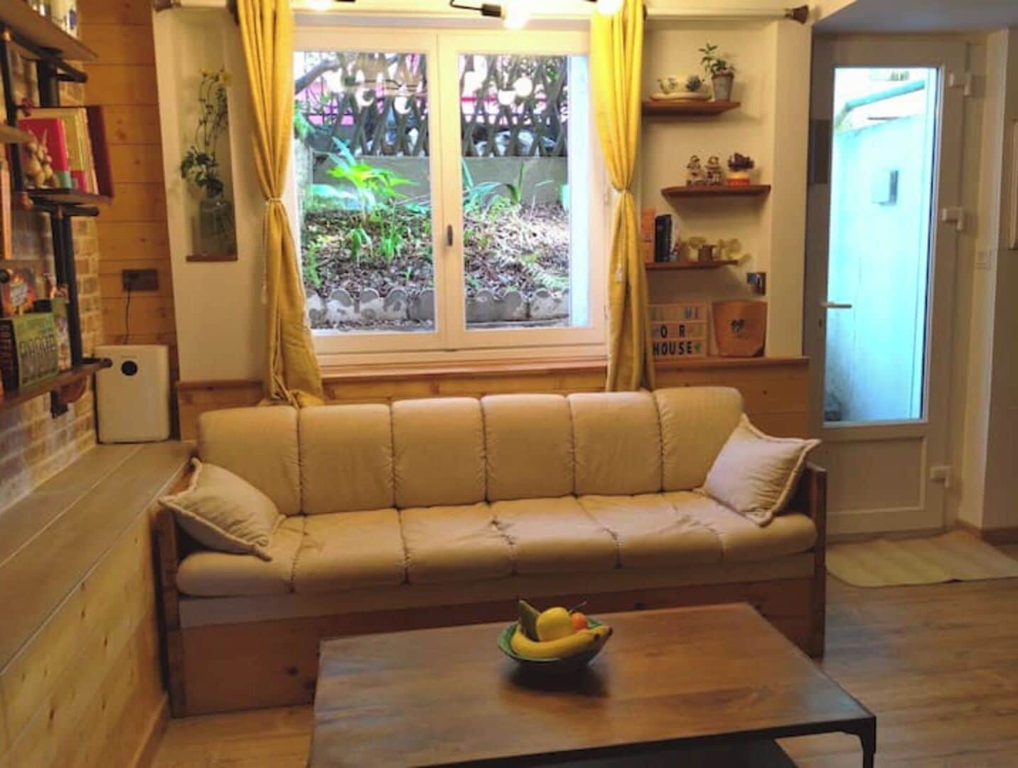 Un salon chaleureux avec un canapé beige, une table basse en bois sur laquelle est posé un panier de fruits, et une fenêtre agrémentée de rideaux.
