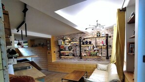 A cozy living room with a wooden sofa, a dining table, and a kitchen area with brick walls and shelves. - Independent industriel chalet @O&R's (Villejuif)