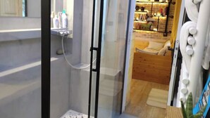 A glass-enclosed shower with a handheld showerhead, a small shelf with toiletries, and a view into a room with a bed and a wooden headboard. - Independent industriel chalet @O&R's (Villejuif)