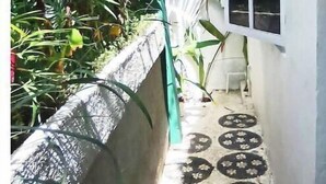 Un étroit couloir extérieur, avec un mur en béton d’un côté et un mur blanc de l’autre. Une fenêtre à motifs et une chaise sont placés contre le mur blanc. Le sol est recouvert de carreaux décoratifs.