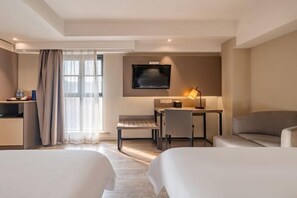 A hotel room with two beds, a desk with a chair, a TV mounted on the wall, and a lamp.