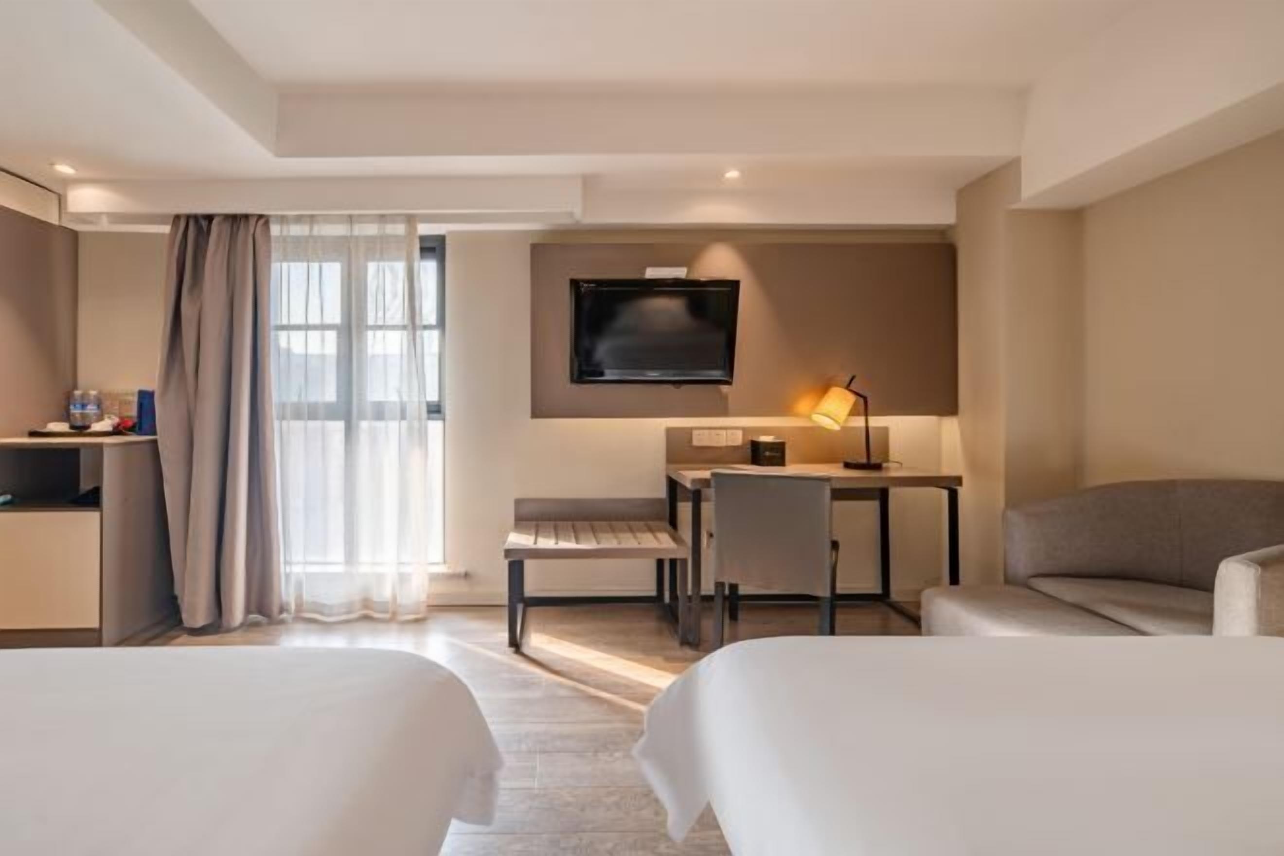 A hotel room with two beds, a desk, a chair, a TV, and a lamp.