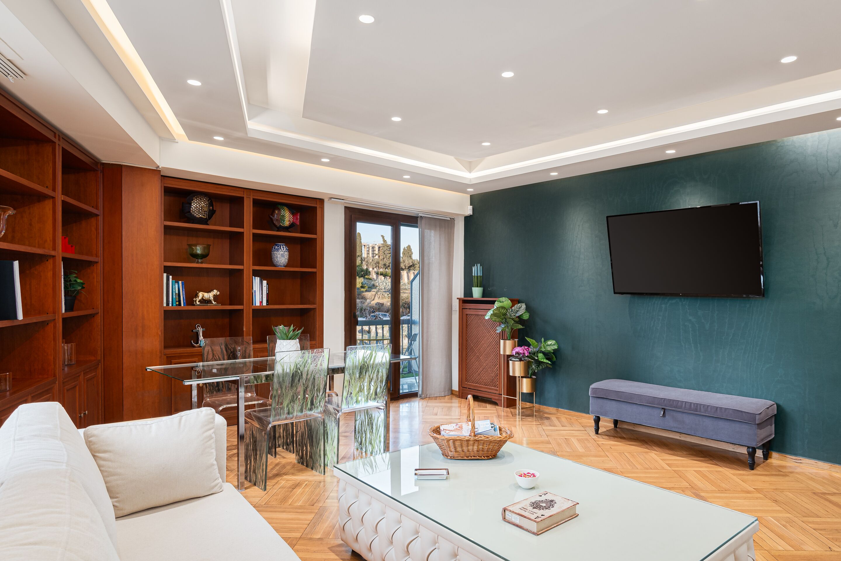 A modern living room with a glass coffee table, white sofa, wooden shelving, and a flat-screen TV mounted on a teal wall.