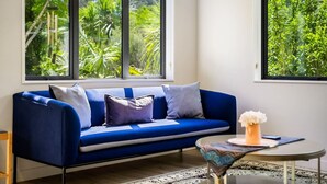A modern living room with a blue sofa, two windows with a view of greenery, and a small table with a vase.