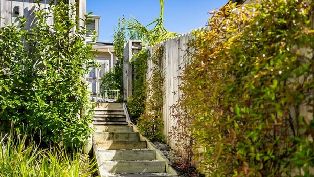 A narrow pathway flanked by lush greenery and a wooden fence, leading to a building with a visible staircase.