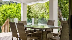 A covered outdoor patio with a glass table and wicker chairs, surrounded by lush greenery.