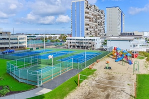 Una cancha polideportiva con canchas de baloncesto y tenis, rodeada de edificios y un parque infantil.