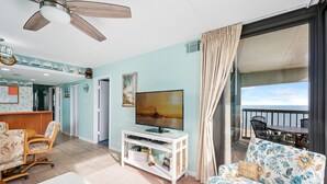A living room with a TV, a sofa, a dining area, and a balcony with a view of the sea. - Beautifully updated 2 bedroom condo by Seaside Vacations & Sales (Ocean City)
