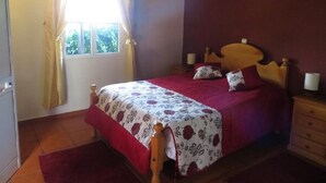 A bedroom with a wooden bed, a window with curtains, a bedside table with a lamp, and a red rug. - Quinta da Quebrada (Santana)