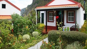 A small house with a red roof and white walls, surrounded by lush gardens and a mountainous backdrop. - Quinta da Quebrada (Santana)