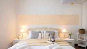 Een netjes opgemaakt bed met witte lakens en twee opgerolde handdoeken, een nachtkastje met een lamp en een kleine vaas, en een bijzettafeltje met een lamp en een decoratief object.