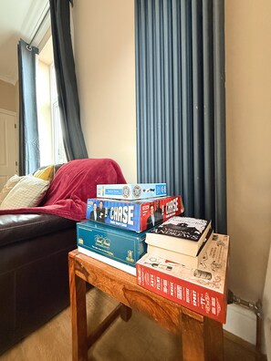 A room with a wooden coffee table holding board games and books, a dark leather sofa with a red blanket, and a window with curtains.