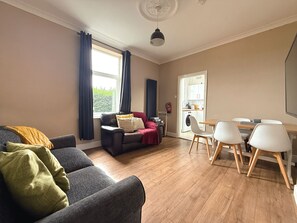 A modern living room with a sofa, a dining table with chairs, a kitchen area, and a large window with curtains. - Spacious & Homely, 4 BR, Parking, Close to Centre (Lowfield)