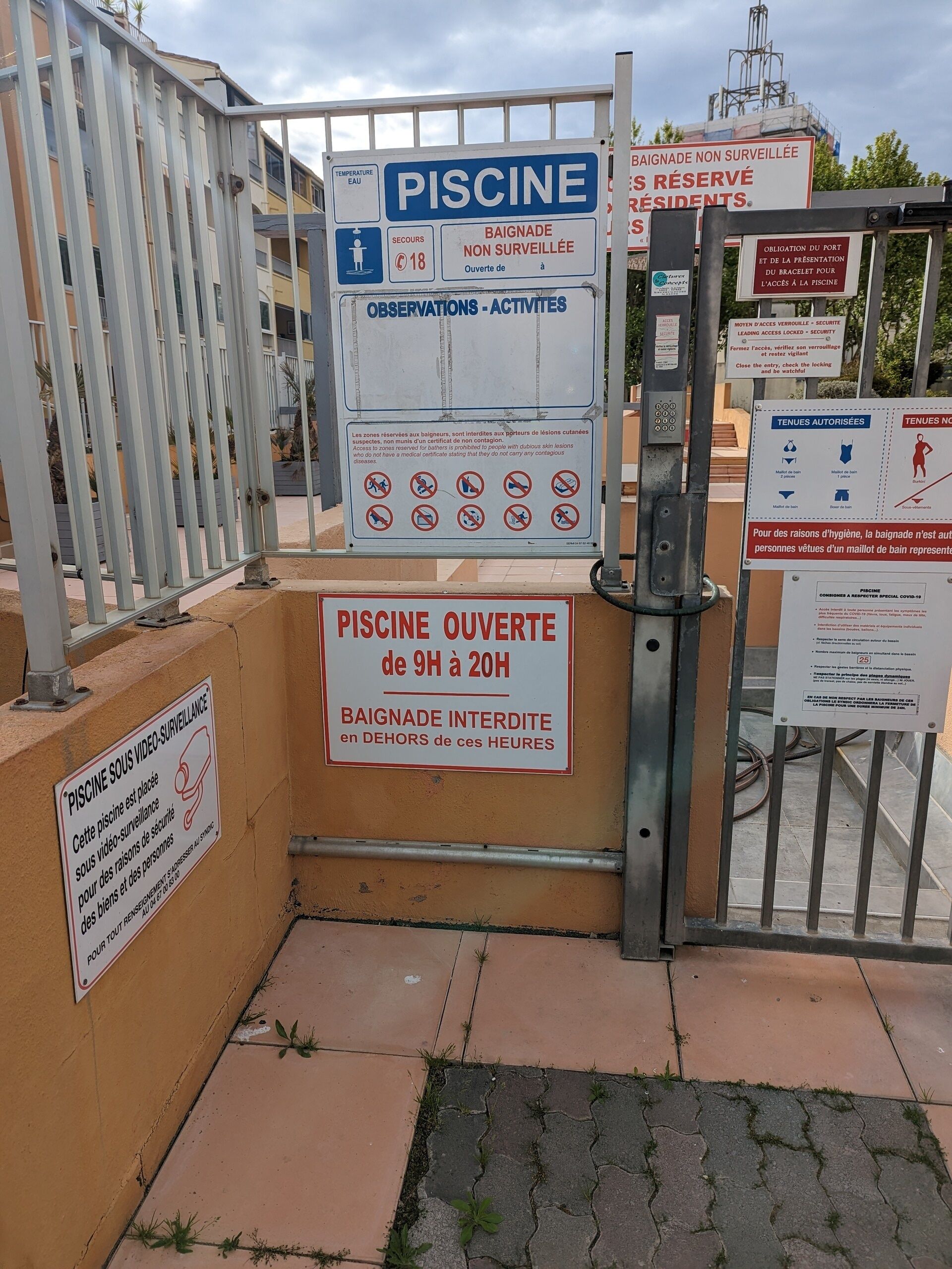 Un espace piscine clos, agrémenté de nombreux panneaux indiquant les règles et les consignes, notamment le prix d’entrée et un avis précisant que la piscine est fermée de 9 h à 20 h.