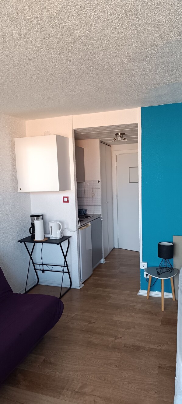 A compact living space with a kitchenette, a purple sofa, a small table with a lamp, and a blue accent wall.