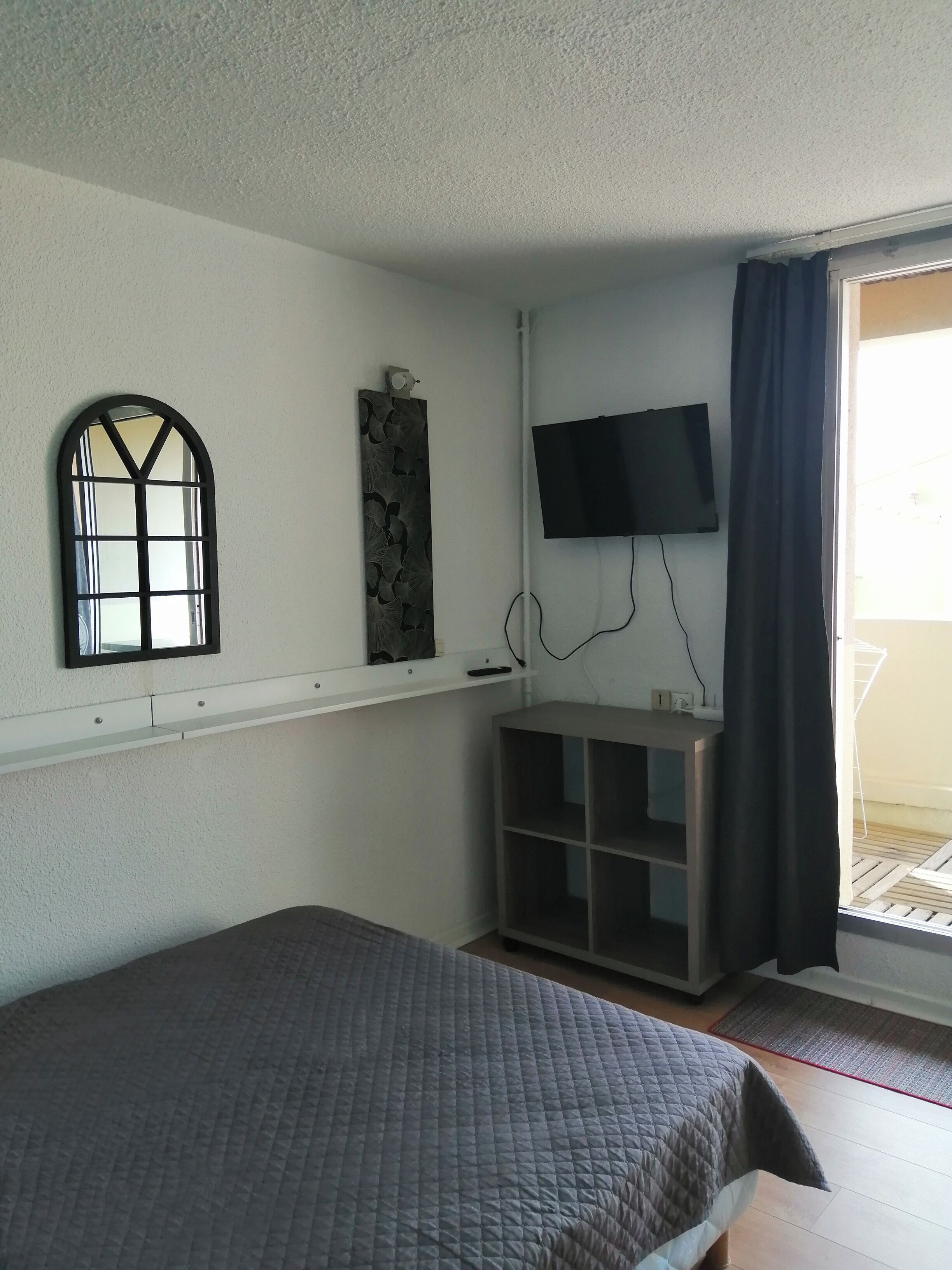 Ein Zimmer mit einem Bett, einem an der Wand befestigten Fernseher, einem Fenster mit dekorativem Rahmen und einem teilweise geöffneten Vorhang.
